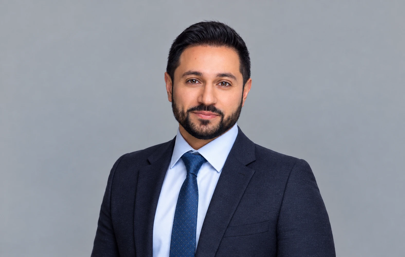 Dr Maytham Omran in a navy suit and pale blue shirt, photographed against a grey studio backdrop